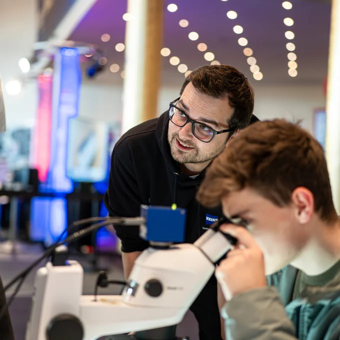 An Image of an \"A Heart for Science\" volunteer helping a young boy using a ZEISS microscope to discover structures of a microchip.\n