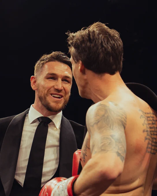 A boxer with tattoos converses with a suited man in a boxing ring environment. The mood is friendly and engaged.