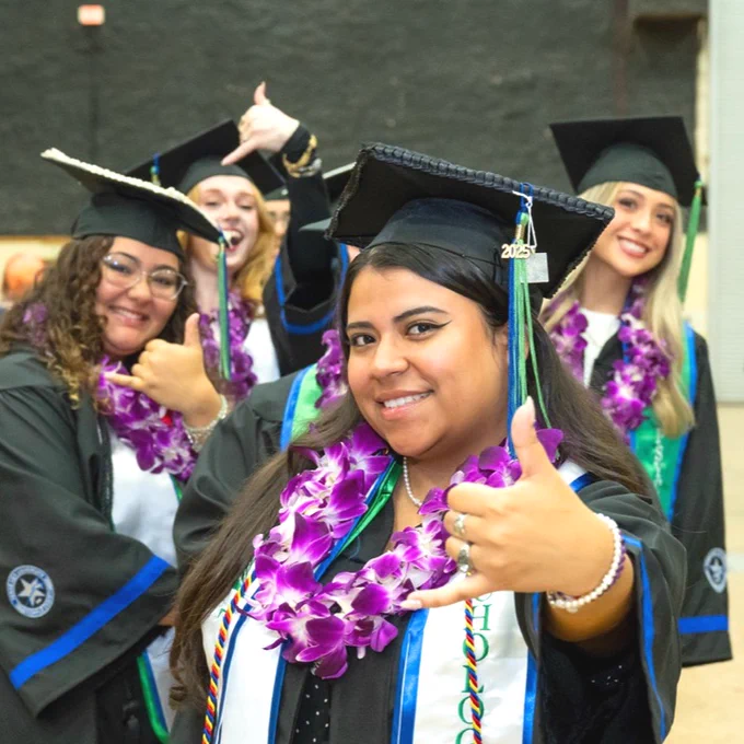 A group of Texas A&M University-Corpus Christi graduates in graduation attire, wearing black gowns and caps adorned with purple leis, smiling and making a shakas up hand gesture.