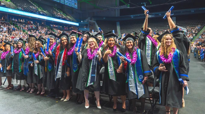 A group of Texas A&M University-Corpus Christi graduates in caps and gowns, adorned with purple leis, stand in an indoor arena celebrating. Many hold rolled diplomas and raise their hands in a shakas up gesture. The background shows a large seated audience.