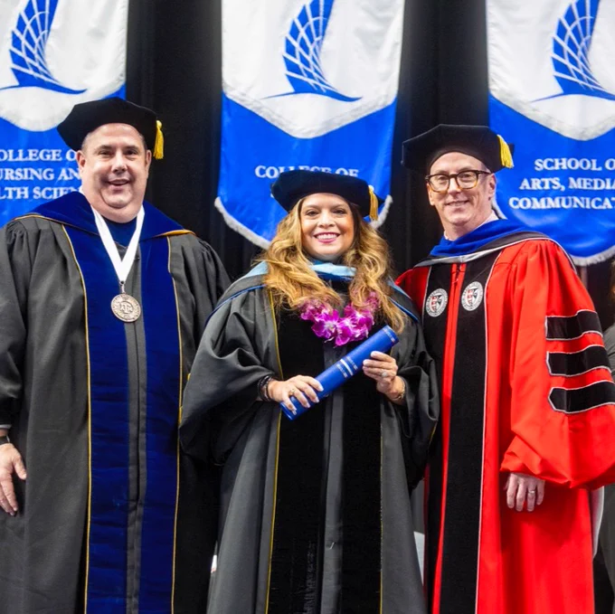 Three individuals in academic regalia stand together at a graduation ceremony. The Texas A&M University-Corpus Christi doctoral graduate in the center is holding a diploma and wearing a lei. Blue and white banners for the College of Nursing and Health Sciences and the School of Arts, Media, and Communication are visible in the background.
