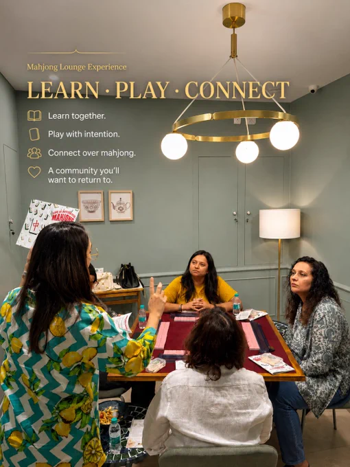 A group of women seated around a Mahjong table during a learning session at Mahjong Lounge in Koramangala, Bangalore. The image features warm olive-green interiors, luxury brass pendant lighting, soft ambient lamps, and a cozy community-focused atmosphere. One participant gestures while teaching or explaining Mahjong strategy as others listen attentively around a red Mahjong table with neatly arranged tiles and learning sheets. Elegant text overlay reads “Learn • Play • Connect” alongside phrases such as “Learn together,” “Play with intention,” and “Connect over Mahjong.” The image captures Mahjong Lounge as a welcoming beginner-friendly Mahjong community space in Bangalore focused on social connection, strategy, mindful play, and offline experiences. The overall mood feels calm, elegant, and community-driven, highlighting women-led game nights, Mahjong classes, and thoughtful social gatherings.
