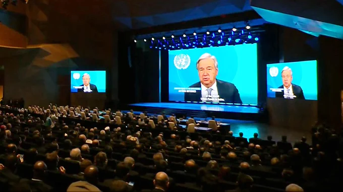 A large conference hall filled with seated attendees faces a stage. Three big screens show António Guterres,  Secretary-General of the United Nations delivering remarks against a blue UN backdrop. Bright stage lights illuminate the scene, and rows of chairs extend across the dimly lit audience area.