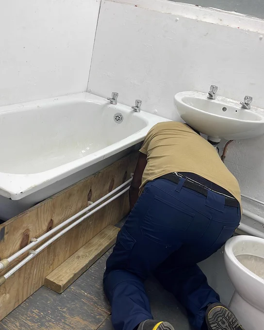 Plumber kneeling on the floor fixing pipes beneath a bathtub in a small bathroom with white walls.