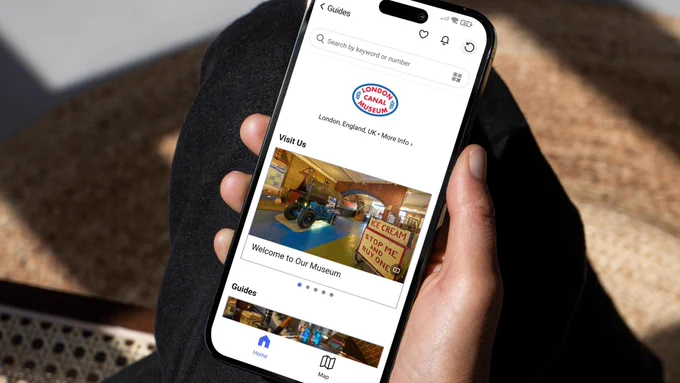 A person holding a smartphone displaying the London Canal Museum guide on the Bloomberg Connects app. The screen shows the museum logo and a "Welcome to Our Museum" section with a photo of a vintage ice cream van.