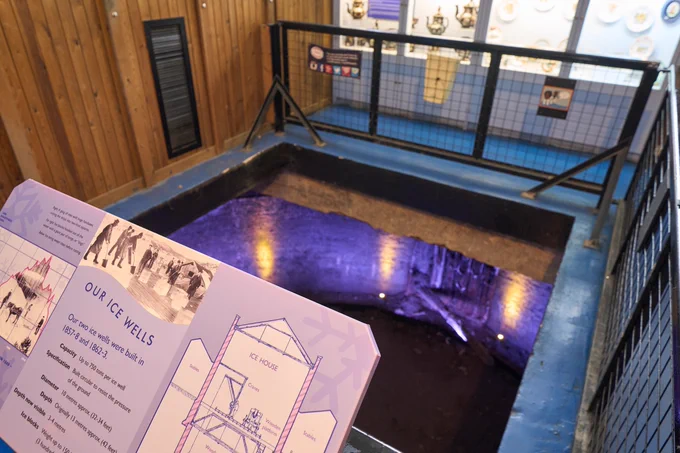 A high-angle view looking down into a large, rectangular opening in the wooden floor of the London Canal Museum. Below the floor level, a deep, circular brick-lined Victorian ice well is visible, illuminated by purple and blue artificial lighting. In the foreground, an educational display board titled 'Our Ice Wells' shows historical diagrams and facts about the 19th-century ice trade.