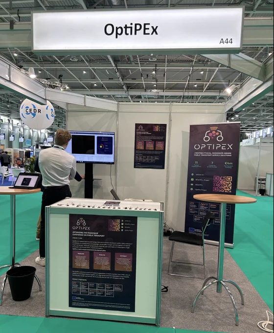 Image shows a stand for the OptiPEx Project at an exhibition. The stand contains two tables, two posters, one screen, one chair and a banner. Two people are looking at the screen. A sign that says 'OptiPEx' is above the stand.