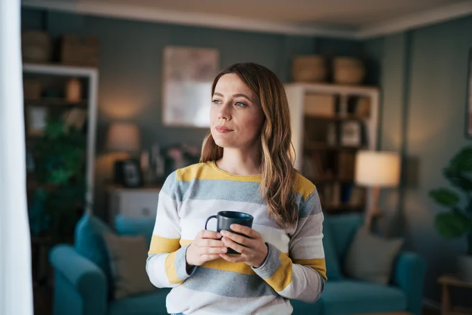 Woman in a striped sweater holding a black mug, standing in a cozy living room with soft lighting and modern decor.