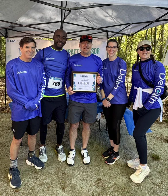 Five members of the Delcath team pose together outdoors at the Boston Miles for Melanoma 5K event hosted by the Melanoma Research Foundation. The group is wearing matching purple Delcath shirts and standing under a sponsor tent, with one team member holding a framed sponsor recognition sign.