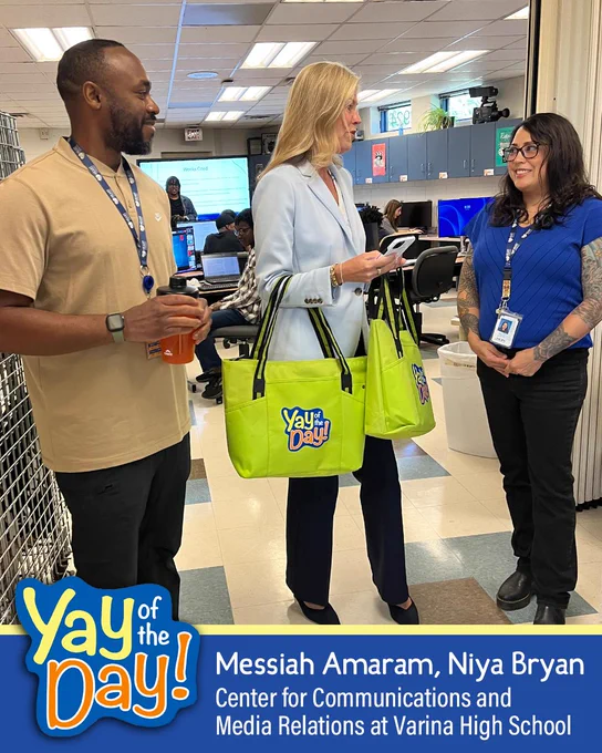 Three people standing in a school hallway, one holding a large green bag with "Yay of the Day!" printed on it.