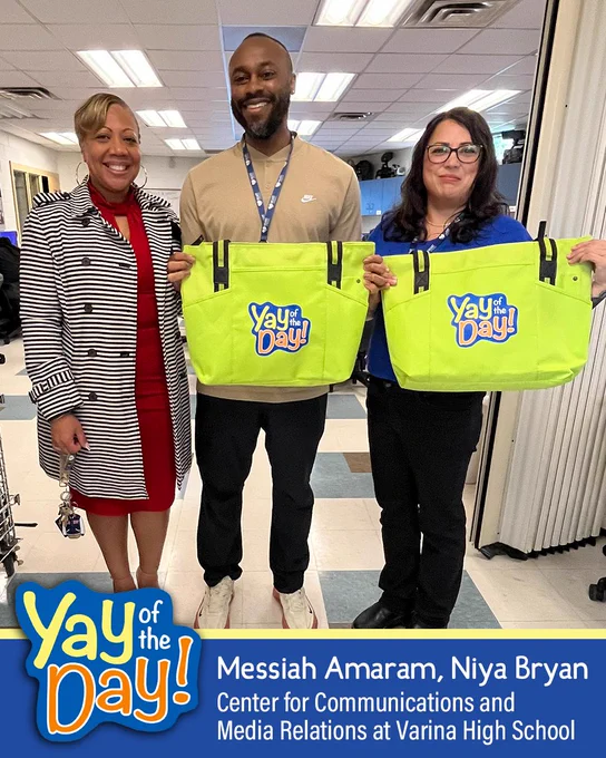 Three people holding bright green tote bags with "Yay of the day!" text, in front of a backdrop with "Varina High School" signage.