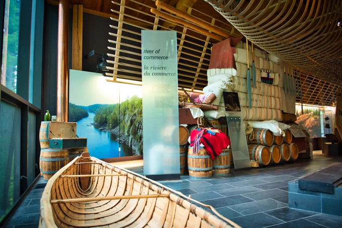 A museum exhibit showcasing a historical display of a fur trading voyageur canoe, large barrels, and colorful trade items. The background features scenic photographs of a river and forest. Signage reads "River of Commerce / La rivière du commerce.
