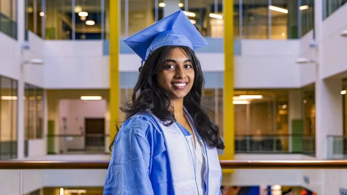 A person in a blue graduation cap and gown smiles indoors, standing in a building atrium with large windows and yellow accents.