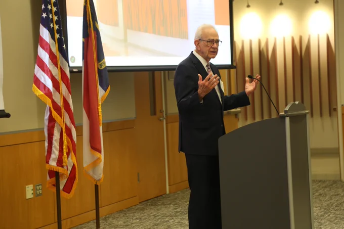 A man in a suit delivers a speech at a podium in a conference room with American and state flags beside him.