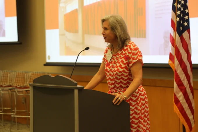 Woman in a red and white patterned dress speaking at a podium with a microphone in a conference room.