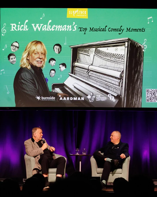 Rick Wakeman and Robert Ross sat in conversation, with the poster for Rick Wakeman’s Top Comedy Moments above.
