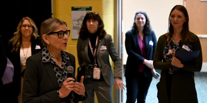 Woman in business attire speaking into a microphone during a professional event with attendees listening in the background.