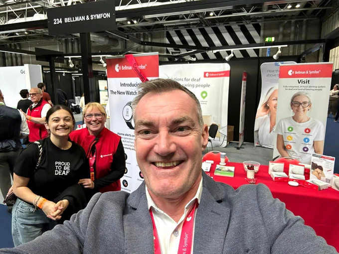 A man in a grey suit holds the camera up to take a selfie. He is smiling at the camera with a Bellman and Symfon branded convention stall behind him, as well as two smiling, female colleagues. 