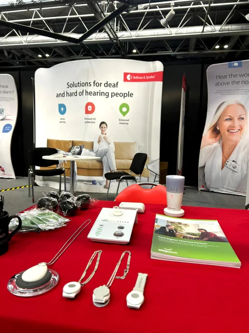 A red cloth-covered table with a display of assistive technology laid out neatly, including Chiptech telecare devices. There is a Bellman and Symfon branded stand in the background.