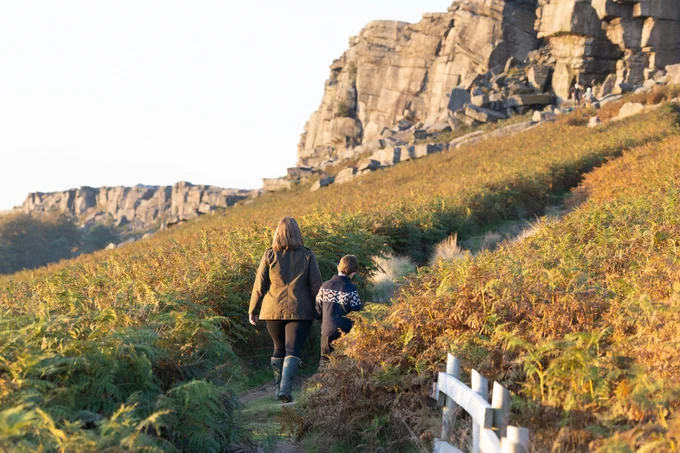 Photo of Stanage Edge at the Peak District. A women and a young boy are walking towards the cliff holding hands. 