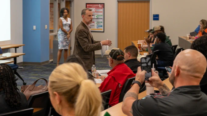 A person stands at the front of a classroom, speaking to an audience seated at tables. The audience is attentive, with a few individuals taking notes and others capturing the moment with their phones. Education materials and water bottles are visible. The room is well-lit with posters on the wall.