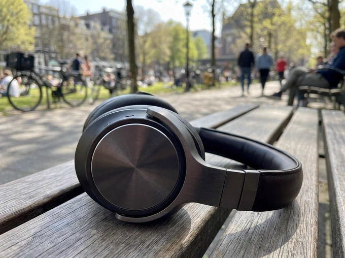A close-up of black headphones sitting on a stone ledge next to a sunny city sidewalk. Abstract, glowing neon wireframe shapes and lines project out from the headphones in shifting colors like blue, pink, green, and purple.
