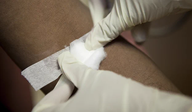 Gloved hands applying a cotton ball and adhesive tape to a person's arm after a blood test.