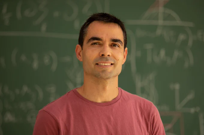 Young man in a red shirt standing in front of a chalkboard filled with mathematical equations and diagrams.