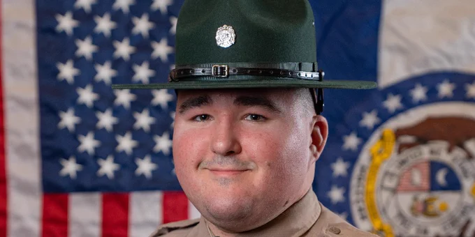 Uniformed park ranger in tan shirt and green hat standing before American and Missouri state flags.