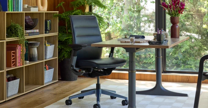 Steelcase Think office chair positioned at a wooden desk with stationery and a vase of flowers by a large window overlooking greenery.