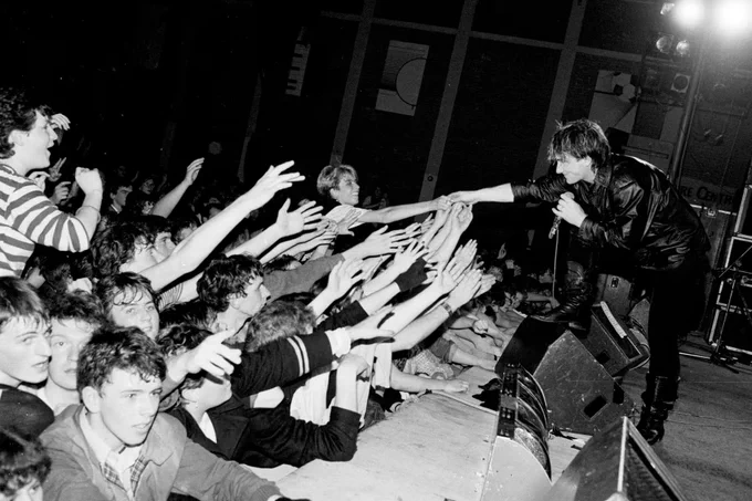 Photo of Bono standing on stage with one foot raised on a monitor, reaching out to the front rows of the audience who are all reaching back to him.