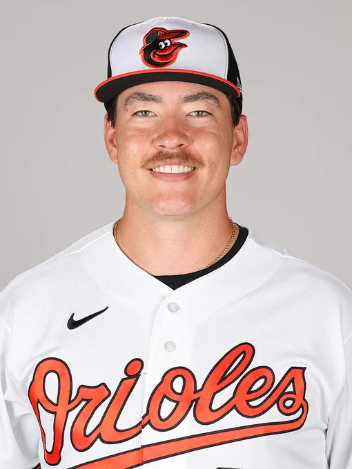 Trey Gibson looks at the camera and smiles. He is wearing a white Orioles jersey and a tri-color cap.