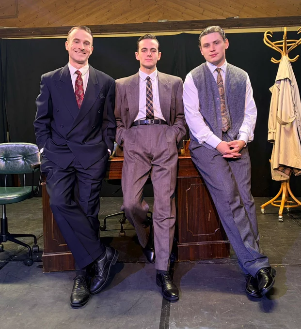 Three white men dressed in 1950s suits and ties resting against a heavy wooden desk on the set of The BBC's First Homosexual.