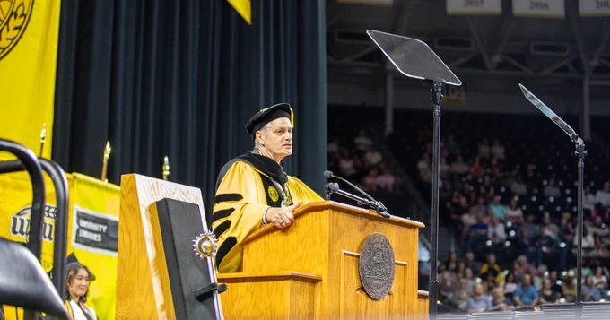 A speaker stands at a podium wearing academic regalia during a commencement ceremony. 