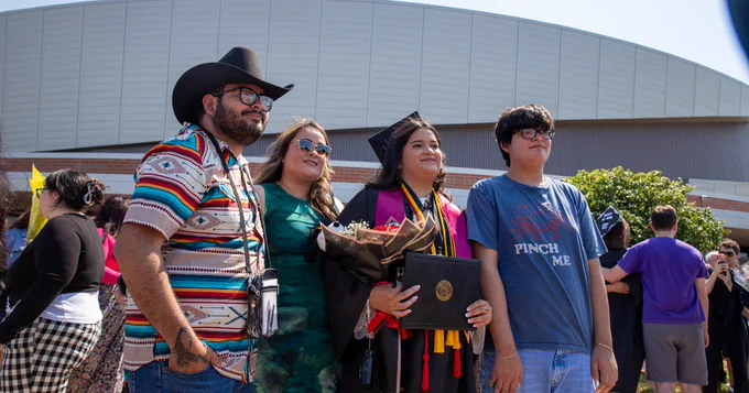 A group of four people stand outdoors, posing for a photo in front of a large building. One person is wearing a graduation cap and gown and holding a diploma and flower arrangement.