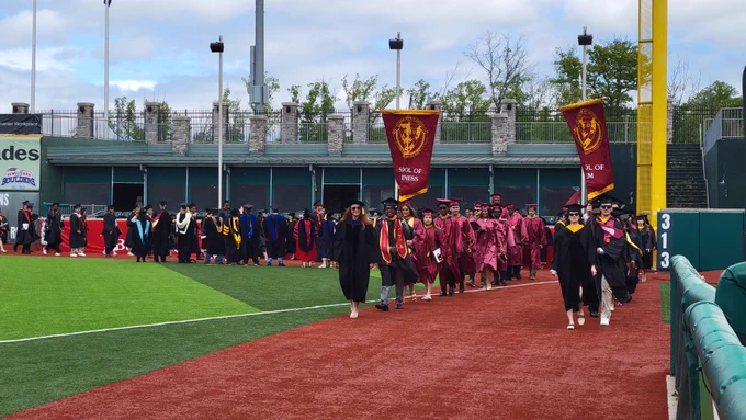 students and faculty processing on field to seats with two gonfalons being carried by students in Schools of Business and STEM
