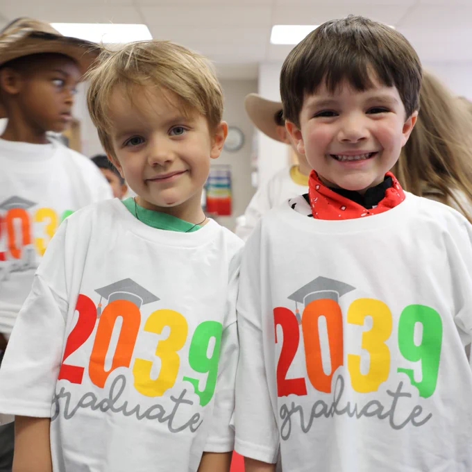 Two children are smiling and wearing white t-shirts with "2039 graduate" printed in colorful text. They are at a school event surrounded by other children.