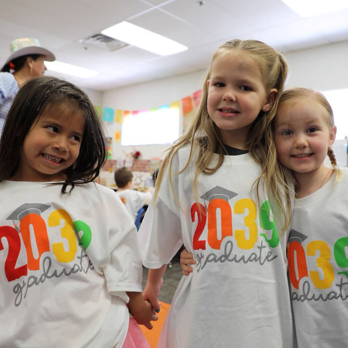 Three children in a classroom wear matching white shirts with "2039 graduate" printed on them in colorful letters. They are smiling and standing close together.