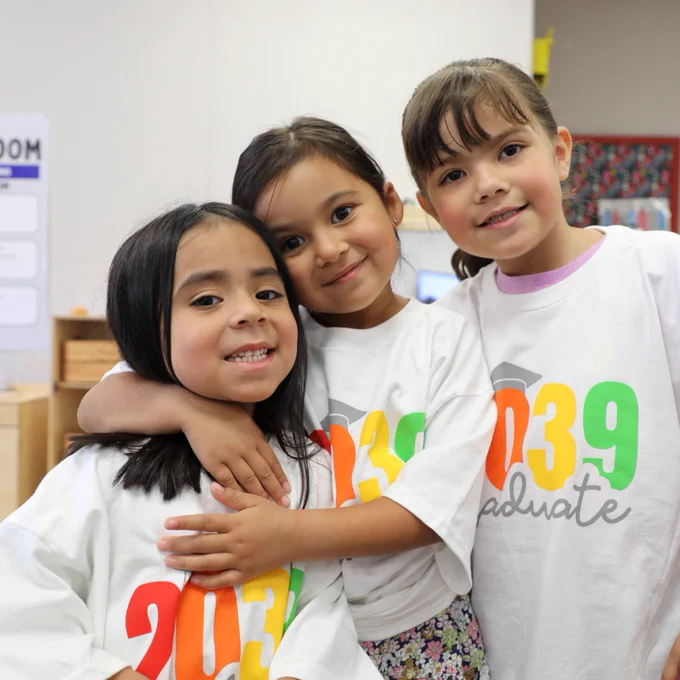 Three children are wearing matching "2039 Graduate" t-shirts stand together, smiling and hugging. The room behind them has classroom elements like charts and colorful displays.