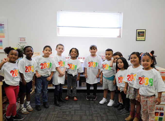 Children are standing in a classroom wearing white T-shirts with "2039 Grad" written on them in colorful letters. They are smiling and standing in a line. The classroom has a bright window and educational posters on the walls.