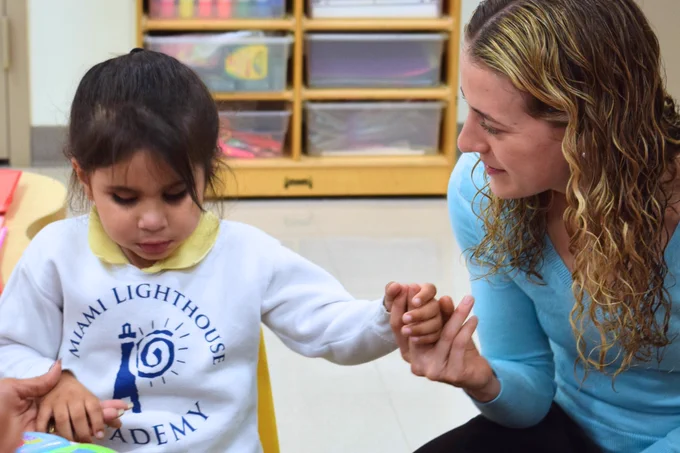 (Photo) A toddler in class at Miami Lighthouse Academy, holding the hand of a visiting doctor. 