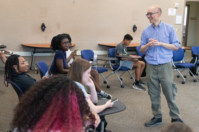 Students sit in a classroom as an instructor stands and leads a discussion, with several students listening and using phones.