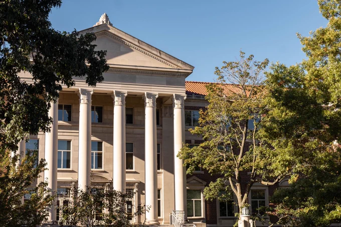 A large, classical building with tall white columns stands surrounded by trees under a clear blue sky.