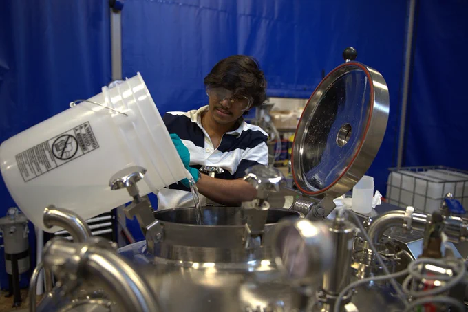 Person wearing gloves pouring liquid from a white bucket into a large stainless steel industrial tank with an open lid