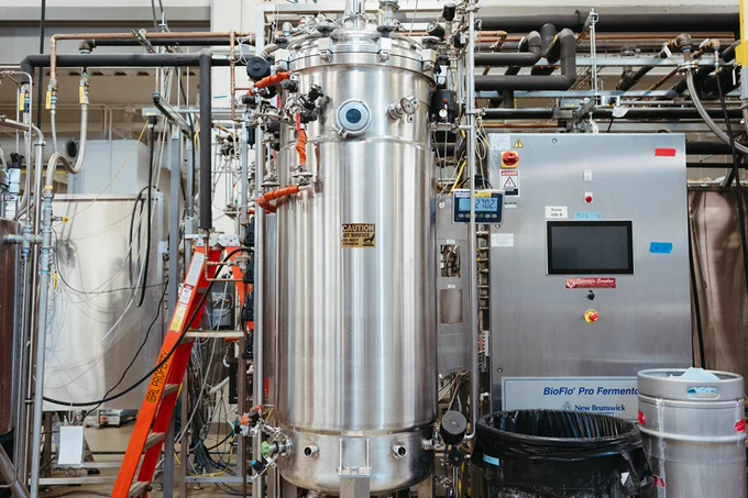 Tall stainless steel fermentation tank with attached pipes and gauges in an industrial lab setting