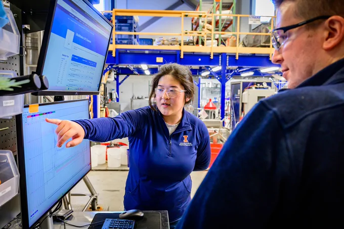 Woman in blue work uniform pointing at data on a large monitor in an industrial workspace with machinery in the background