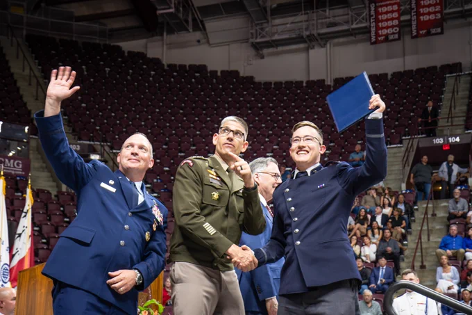 Three uniformed officials stand on stage in front of arena seating; one points upward while another graduate in a blue uniform raises a diploma cover, and they shake hands during the ceremony. 