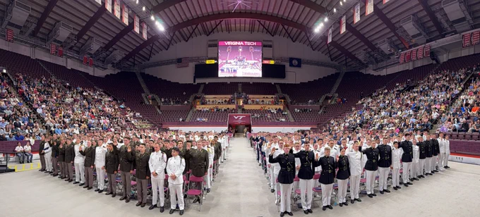 Wide panoramic view inside Virginia Tech’s Cassell Coliseum during a Corps of Cadets graduation ceremony, with rows of uniformed cadets standing and raising their right hands while a large audience fills the surrounding maroon stadium seating beneath a scoreboard reading “Virginia Tech.”