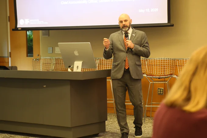 Man in a suit giving a presentation with a microphone in front of a large screen and a laptop on a podium.