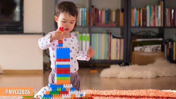 Young child building a tall colorful block tower on a carpeted floor in a cozy living room with bookshelves in the background.